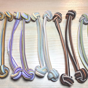 May include: A collection of colorful hair ties with knotted ends. The elastic bands feature various striped patterns in shades of purple, yellow, orange, and brown. The knotted ends are also multi-colored.