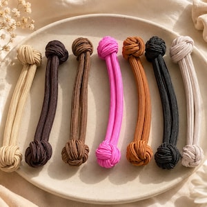 May include: A set of seven fabric hair ties, each featuring a long, ribbed cord with a decorative knot at both ends. The colours include beige, dark brown, light brown, bright pink, orange-brown, black, and off-white, arranged in a row.