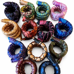 May include: A collection of twelve circular fabric-wrapped hair accessories arranged in a grid. Each ring features a coiled design with contrasting thread lacing, showcasing various colors including black, gold, green, red, pink, and blue.