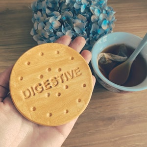 Op de afbeelding: Een ronde, bruinbeige keramische onderzetter met het woord "DIGESTIVE" in reliëf. De onderzetter heeft kleine, gelijkmatig verdeelde gaatjes langs de rand, die op een koekje lijken. Een kopje thee en blauwe bloemen op de achtergrond.