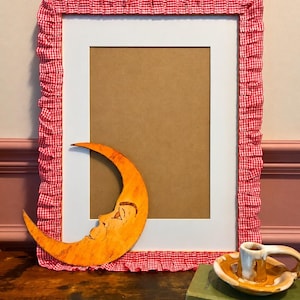 May include: A rectangular picture frame featuring a red and white gingham fabric ruffle border. An orange crescent moon cutout with a hand-drawn face rests against the frame. A small ceramic candle holder sits on a green book titled Wild Flowers.