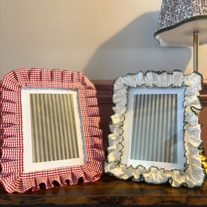 May include: Two rectangular picture frames with fabric borders. The left frame features a red and white gingham fabric ruffle. The right frame features a cream and grey striped fabric ruffle with a dark green edge. Both frames hold grey striped inserts.