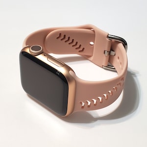 May include: A rose gold Apple Watch with a light pink silicone band featuring a perforated design. The band has a silver buckle.