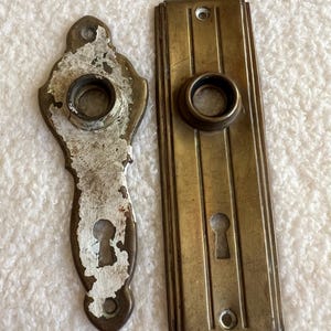 May include: Two antique brass door hardware pieces. One is a rectangular plate with a keyhole and a round hole. The other is a decorative piece with a keyhole and a round hole. Both pieces have a worn, aged look.