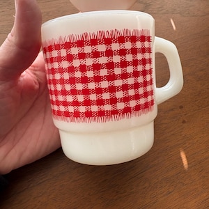 May include: A white ceramic mug with a red and white checkered pattern. The mug has a handle and a slightly flared base. A vintage-style decorative head is in the background.