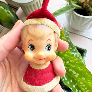 May include: A vintage Christmas elf ornament with a red felt hat and tunic, and a painted face with blue eyes and rosy cheeks. The elf has a smiling expression and is a classic holiday decoration.