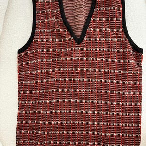 May include: A red, black, and white plaid sweater vest with a V-neck and black trim. The vest has a label that reads "Jones New York". The pattern is a grid of vertical and horizontal lines.