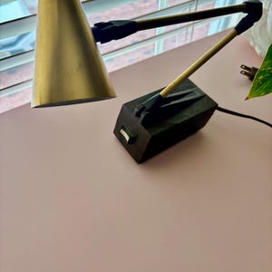 May include: A brass-colored desk lamp with a conical shade and a black, adjustable arm. The lamp sits on a pink surface. The base of the lamp is a dark, rectangular block.