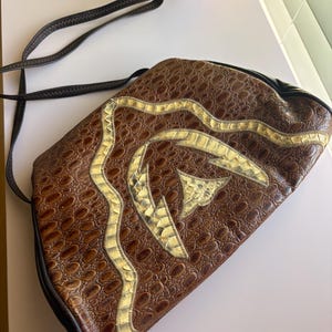 May include: A brown, semi-circular shoulder bag with a textured, embossed pattern resembling reptile skin. The front features a decorative, winding appliqué in a metallic gold, scale-like material. It includes a thin, dark brown shoulder strap.