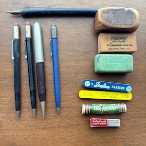 May include: A collection of vintage writing supplies, including pencils, erasers, and lead refills, arranged on a wooden surface. The items include a black dip pen, mechanical pencils, and various erasers with printed labels. The color palette is dominated by browns, blacks, and blues.