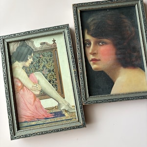May include: Two rectangular framed prints with dark grey, textured borders featuring geometric patterns. One print depicts a person in a pink dress sitting on a patterned chair. The other print shows a portrait of a person with dark hair and rosy cheeks.