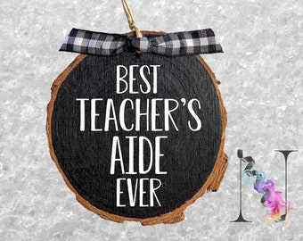 May include: A round wooden ornament with a black background and white text that reads "Best Teacher's Aide Ever". The ornament is decorated with a black and white checkered ribbon.