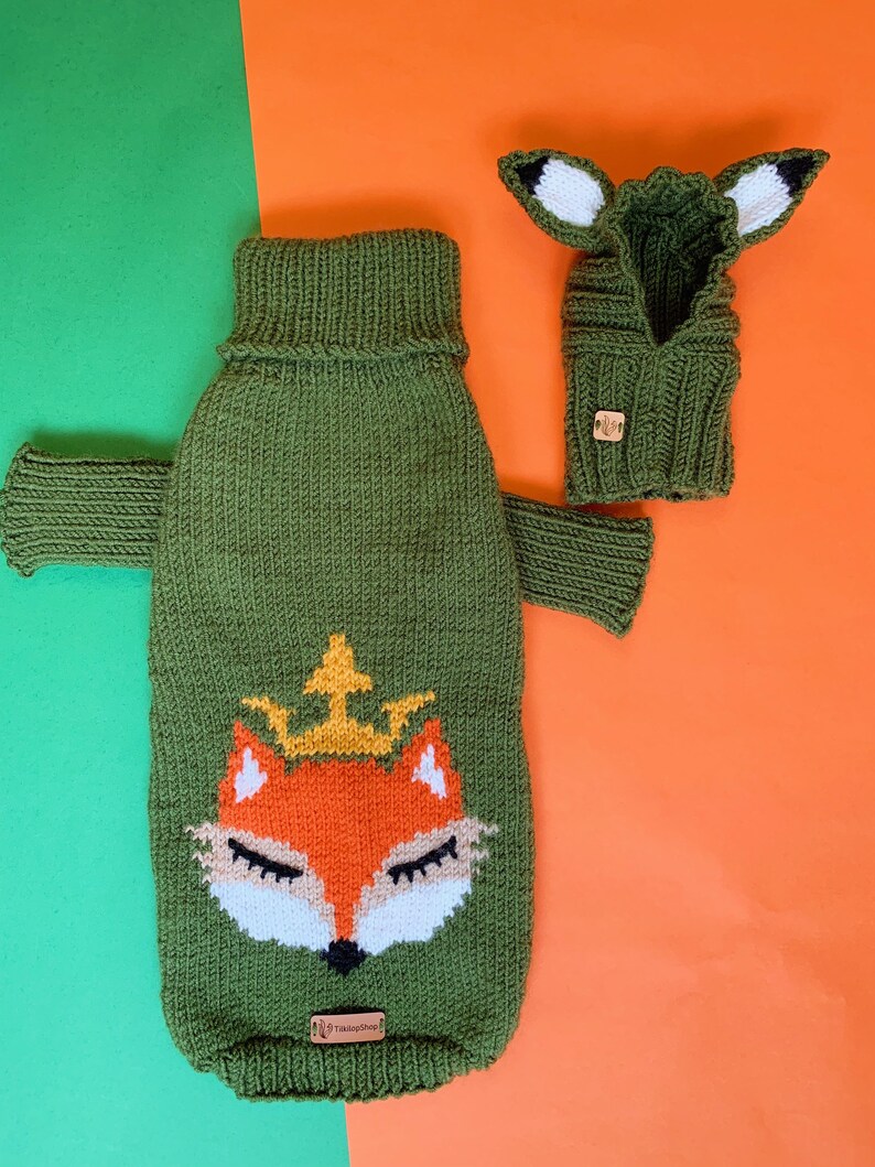 Puede incluir: Su&eacute;ter de punto verde para perro con un dise&ntilde;o de cara de zorro y un gorro de zorro de punto verde a juego. La cara del zorro tiene una corona en la cabeza y es naranja, blanca y negra.