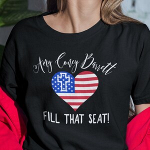 May include: Black t-shirt with a white heart design featuring an American flag and a cross. The text "Amy Coney Barrett" and "Fill That Seat!" are printed on the shirt.