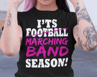 Puede incluir: Una camiseta negra con el texto "It's Football Marching Band Season!" en letras blancas, con la palabra "Football" tachada y las palabras "Marching Band" en letras rosas.