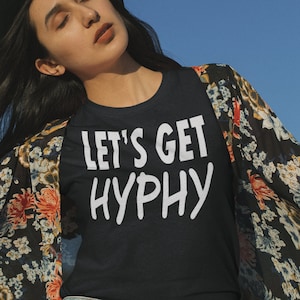 May include: A woman wearing a black t-shirt with the text "Let's Get Hyphy" in white lettering. She is wearing a floral kimono and denim shorts. She is also wearing a black hat.