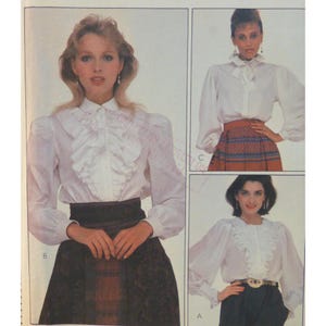 May include: A sewing pattern illustration featuring three variations of a white long-sleeved blouse. The designs include ruffled front plackets, high collars, and gathered sleeve cuffs, paired with dark skirts or trousers in various styles.