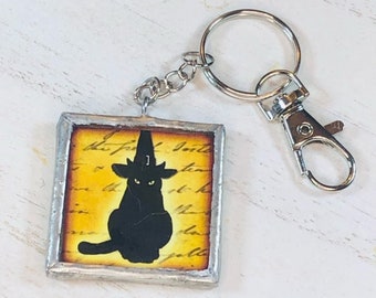 May include: A black cat wearing a witch's hat is featured on a square, silver-framed tile keychain. The tile has a yellow background with black script text. The keychain is attached to a silver split ring and a silver lobster clasp.