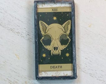 May include: A rectangular pendant featuring a cat skull tarot card design. The card has a dark green background with gold stars and the word "DEATH" at the bottom. The top of the card reads "XIII". The pendant is framed in a dark metal.