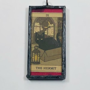 May include: A rectangular pendant featuring a tarot card design. The card depicts a black cat in a box, with a window, moon, and the text "THE HERMIT". The pendant has a black frame with a red border and a metal loop for hanging.