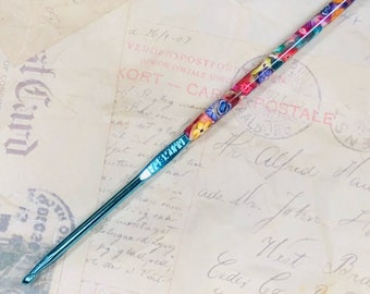 May include: A turquoise crochet hook with a floral design painted on the handle. The hook is made of metal and has a pointed tip. The floral design is colorful and includes pink, red, yellow, and blue flowers.