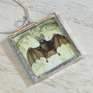 May include: A silver necklace with a square glass pendant featuring a vintage illustration of a brown bat with large ears against a green and white background.