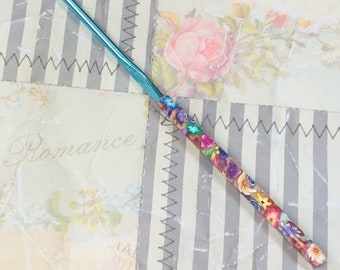 May include: A blue crochet hook with a colorful floral design. The hook is made of metal and has a smooth finish. The floral design is made of polymer clay and is very detailed.