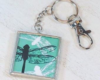 May include: A silver key chain with a square charm featuring a green background with a black dragonfly and white dragonfly silhouettes.