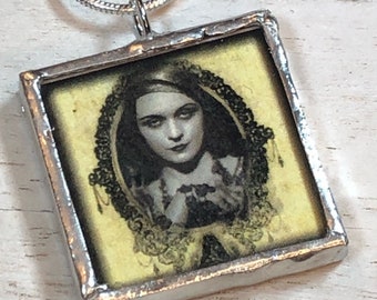May include: A silver necklace with a glass pendant featuring a black and white image of a woman with dark hair and a lace collar. The woman is looking at the viewer with a serious expression.