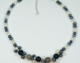 May include: A silver necklace with black, white, and gray pearls. The pearls are arranged in a graduated pattern, with the largest pearls in the center of the necklace. The necklace is accented with small silver beads.