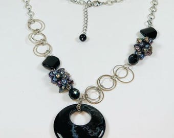 May include: A silver chain necklace with a large black agate pendant. The pendant has a circle cut out of the center. The necklace also features black and iridescent beads and silver rings.
