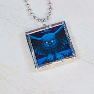 May include: A silver-toned necklace featuring a square pendant with a blue owl illustration. The owl has yellow eyes and outstretched wings against a dark blue background. The pendant is framed with a silver border and hangs from a ball chain.