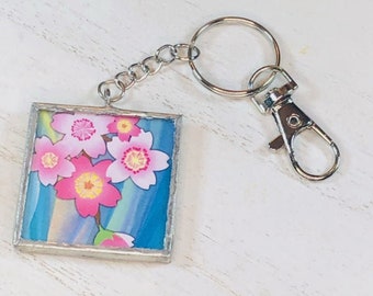 May include: A silver keychain with a square charm featuring a pink and white floral design on a blue background.