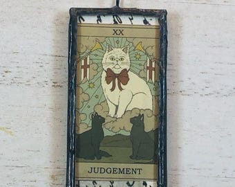 May include: A rectangular stained glass pendant featuring a tarot card design. The card depicts a white cat with a bow tie, two black cats, and the word "JUDGEMENT". The frame is black with a loop for hanging.