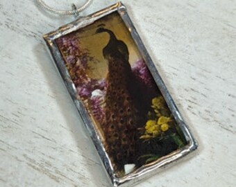 May include: A stained glass pendant featuring a peacock with a detailed pattern of feathers. The peacock is standing in a garden setting with flowers and a moon in the background. The pendant is framed in silver.