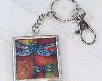 May include: A silver key chain with a square glass charm featuring a colorful dragonfly illustration on a red background.