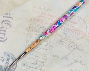 May include: A close-up of a colorful crochet hook with a floral design. The hook is made of metal and has a wooden handle. The hook is lying on a piece of paper with writing on it.