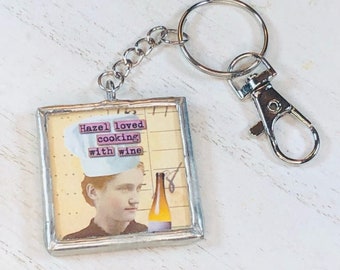 May include: A silver key chain with a small square picture frame charm. The frame has a vintage image of a person wearing a chef's hat and the text "Hazel loved cooking with wine".