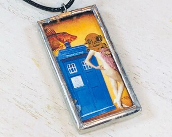 May include: A silver framed pendant necklace with a colorful image of a blue police box, a diver, a fish, and a woman. The image is set against a yellow background.