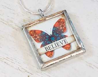 May include: A silver necklace with a glass pendant featuring a colorful butterfly with the word "BELIEVE" on a banner.