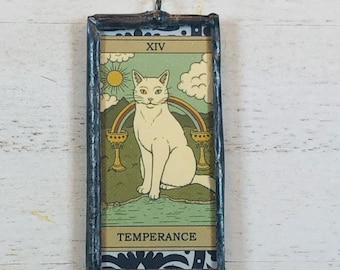 May include: A rectangular, stained glass tarot card featuring a white cat sitting in front of a rainbow, with the text "TEMPERANCE" at the bottom. The card has a black border and a small loop at the top for hanging.