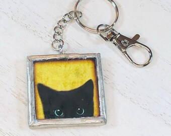 May include: A black cat with green eyes peeks out from behind a yellow background in a silver framed keychain.