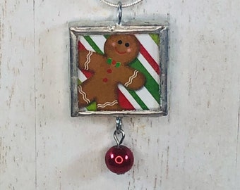 May include: A silver-toned necklace featuring a square pendant with a gingerbread man design. The pendant has a red and green striped background and a small, red, round charm hanging below. The necklace is on a white surface.