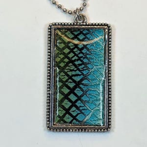 May include: A rectangular pendant necklace with a silver-colored beaded frame. The pendant features a textured pattern in shades of green and blue. The necklace has a silver-colored ball chain.