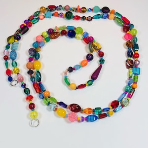 May include: A long multi-strand necklace made of colorful glass beads in various shapes and sizes. The necklace is made of many different colors, including red, orange, yellow, green, blue, purple, and pink.