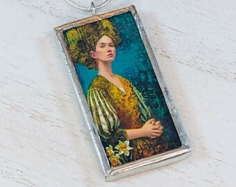 May include: A silver chain necklace with a rectangular pendant featuring a woman with a floral crown, wearing a yellow and white striped dress, against a blue and green background.