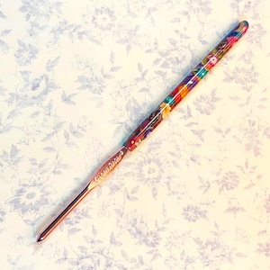 May include: A Susan Bates crochet hook with a colorful floral design on the handle. The hook has a rose gold metal tip and the brand name is printed on the handle.