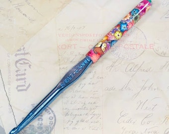 May include: A blue crochet hook with a colorful floral and butterfly design on the handle. The handle is made of polymer clay and features a variety of colors, including pink, yellow, green, and blue. The hook is labeled "Kollage 12" on the metal portion.