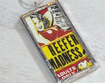 May include: A silver framed glass pendant with a vintage movie poster image. The poster is yellow, red, and black with the text "Reefer Madness Adults Only".
