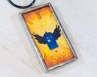 May include: A silver framed glass pendant necklace with a yellow, orange, and red background featuring a blue TARDIS with black wings. The TARDIS is surrounded by a clock face with Roman numerals.